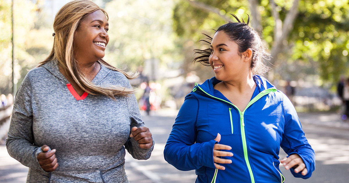 Two young women of color stand next to each other in a park, smiling at each other. Both are wearing athletic clothes.