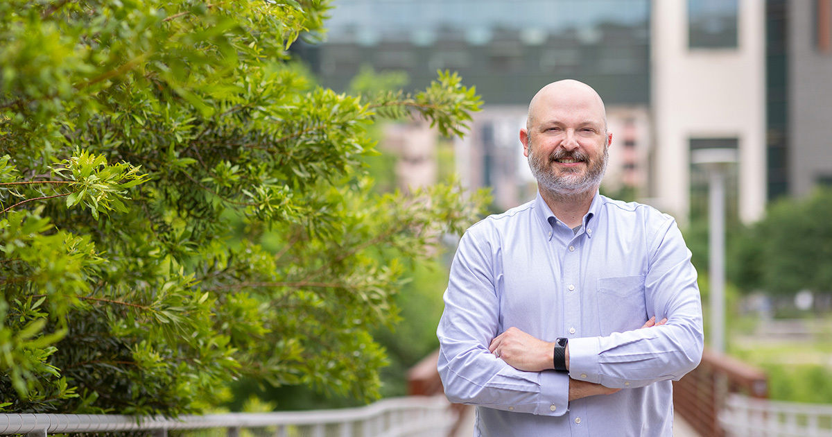 Dr. Jared Benge is standing outside of the HTB on a bridge surrounded by green foliage.  He is crossing his arms, smiling, and wearing a light purple shirt.