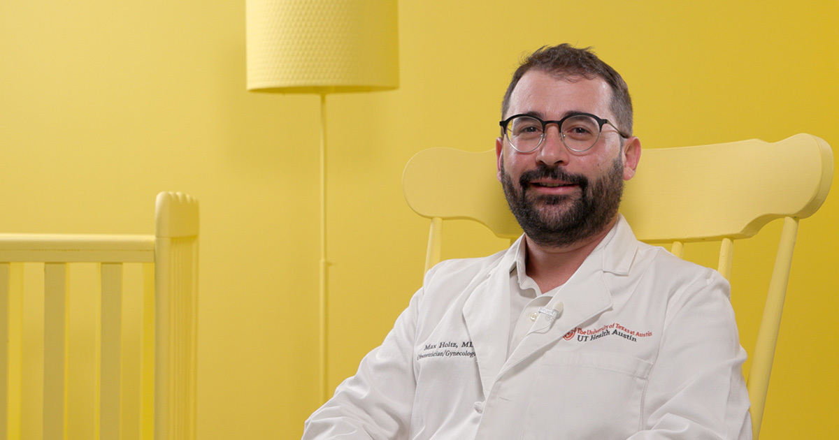 A physician wearing a white coat and glasses sits in a bright yellow rocking chair in a yellow nursery.