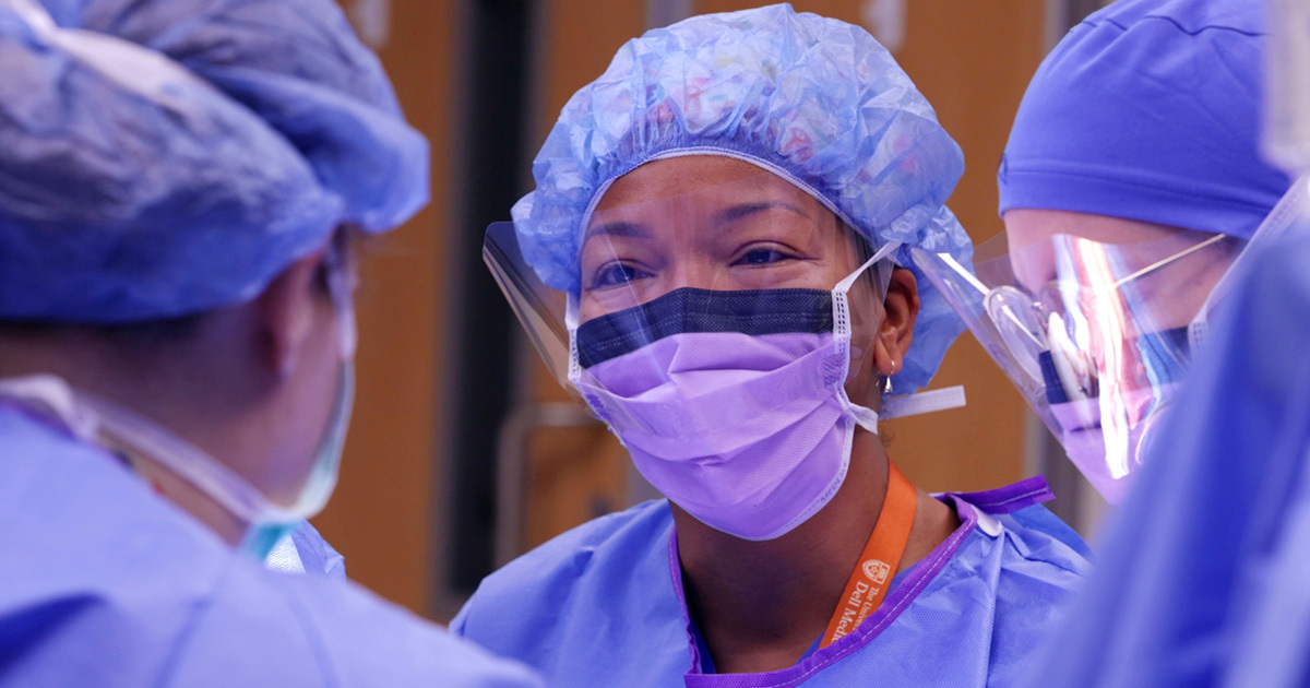 M. Yvette Williams-Brown, MD, wearing a surgical mask, face guard, scrubs, and head covering in the operating room surrounded by colleagues.