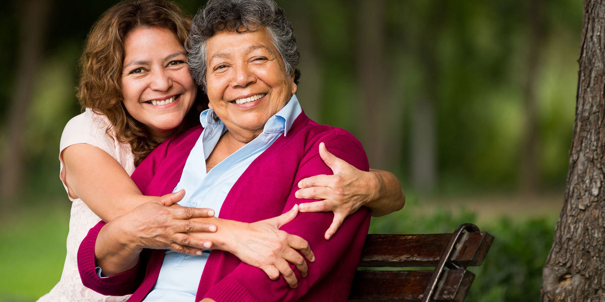 An older Latinx woman sits on a park bench. Her daughter sits next to her with arms around her shoulders. Both face the camera, smiling.