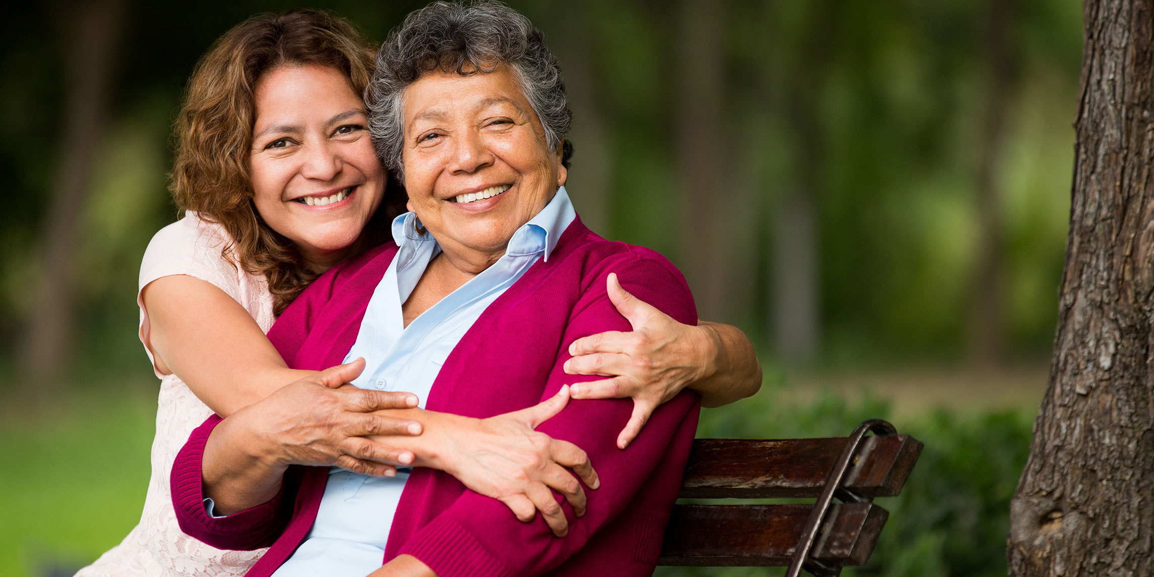 An older Latinx woman sits on a park bench. Her daughter sits next to her with arms around her shoulders. Both face the camera, smiling.