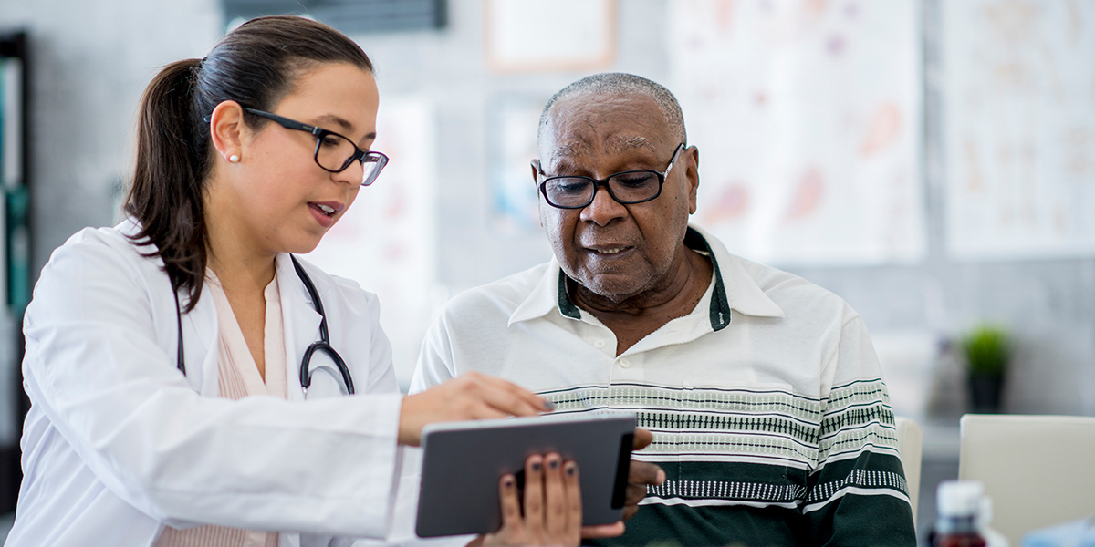 A female clinician points to the screen on a digital tablet device while speaking to an elderly Black man.