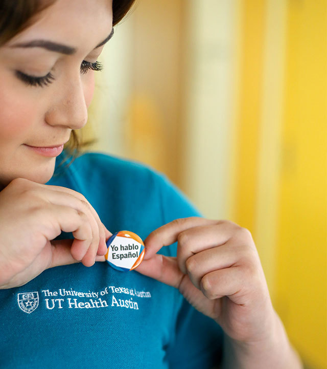 A woman places a button reading "Yo hablo espanol" on her polo shirt. Embroidered on the shirt is the logo of UT Health Austin.