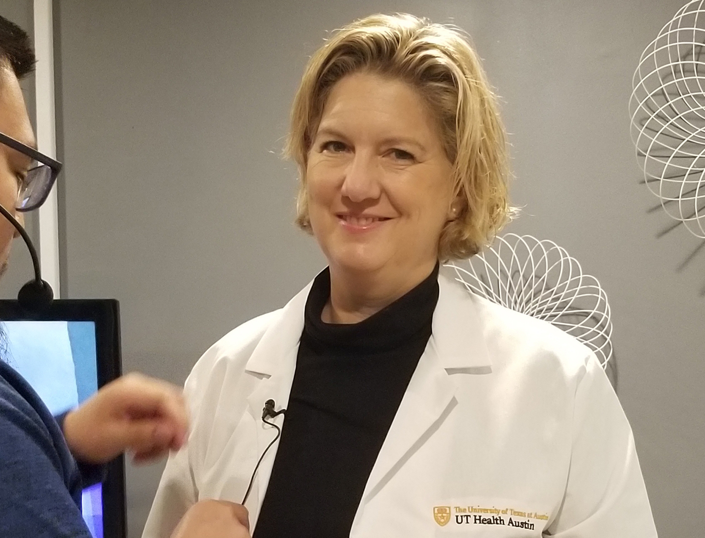 Elizabeth Kvale, MD, Director of Survivorship for the LIVESTRONG Cancer Institutes stands while a person places a lavalier microphone on the lapel of her lab coat.