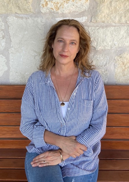 A woman wearing a blue button up shirt sits on wooden bench with her arms in her lap while looking at the camera.