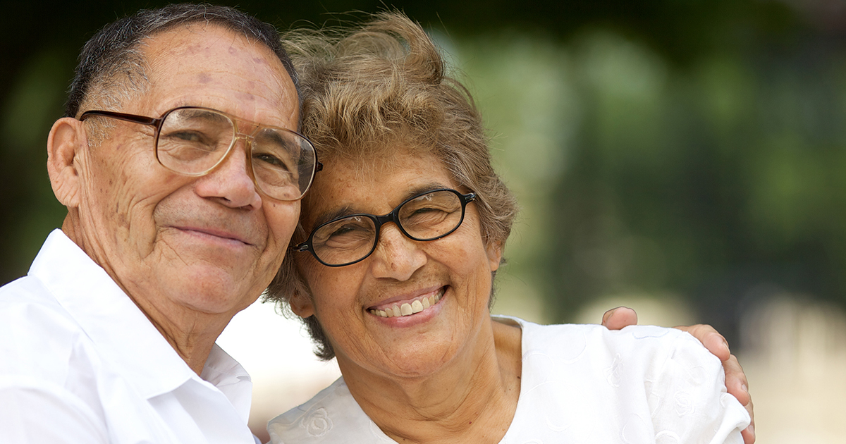 An older Latinx couple sit with his arm around her shoulders, smiling into the camera.