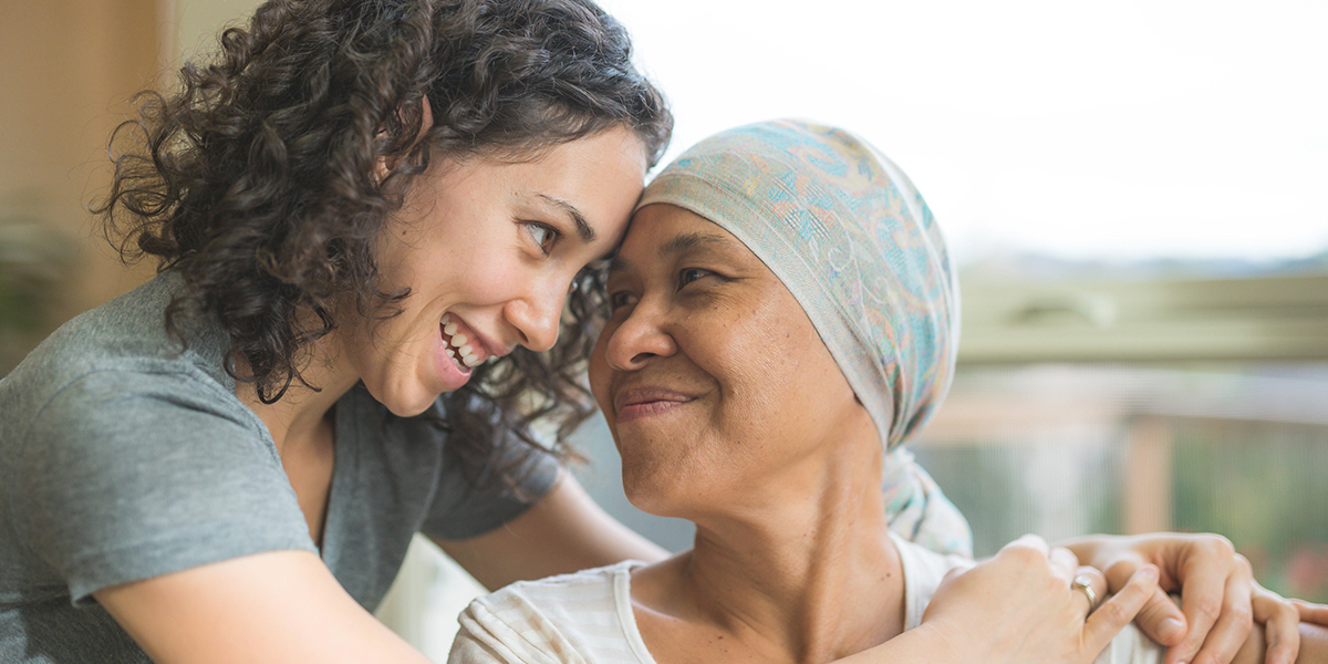 A young woman smiles at and embraces an older woman wearing a headscarf who is seated and looking at the younger woman.