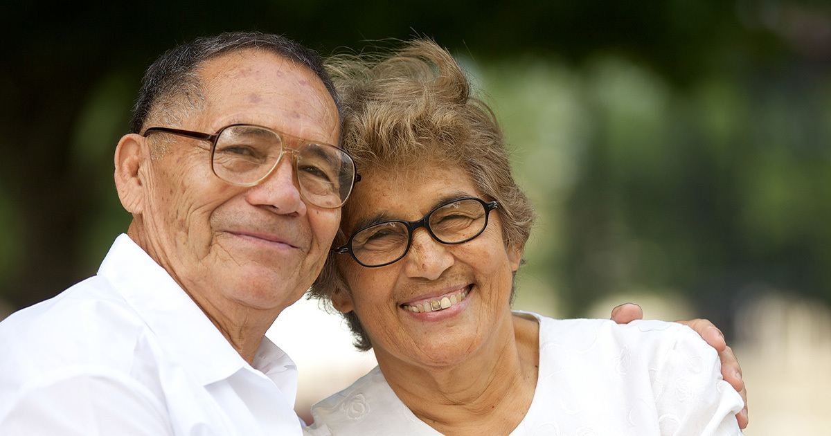 An older LatinX couple are sitting and smiling at the camera on a park bench.