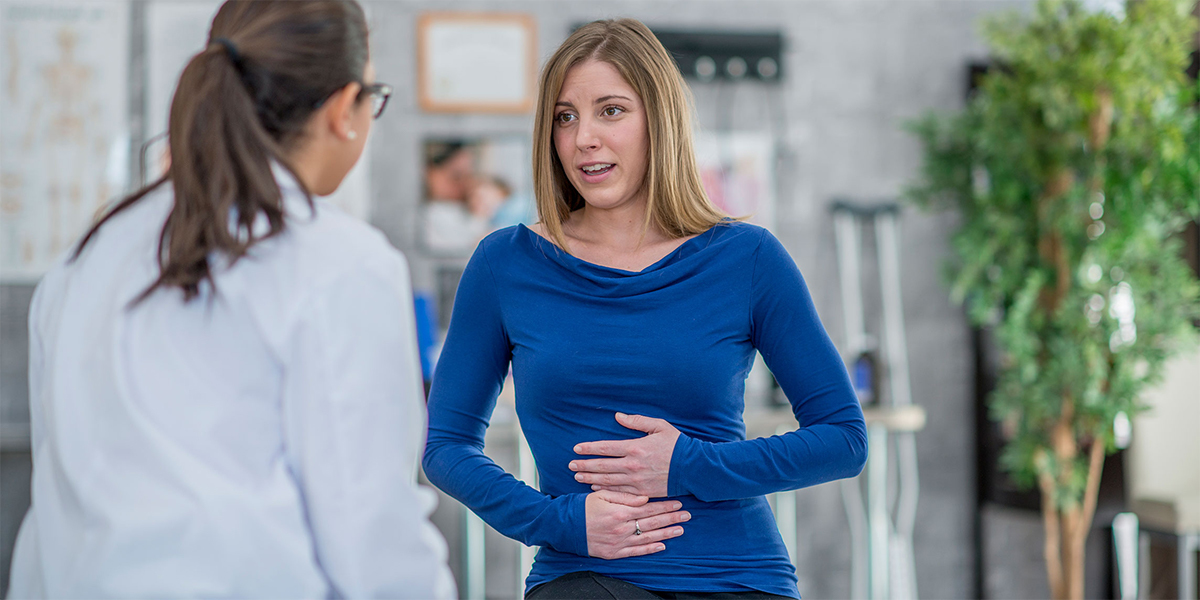 A female clinician, her back turned to the camera, speaks with a woman who holds her hands on her stomach in an expression of discomfort.