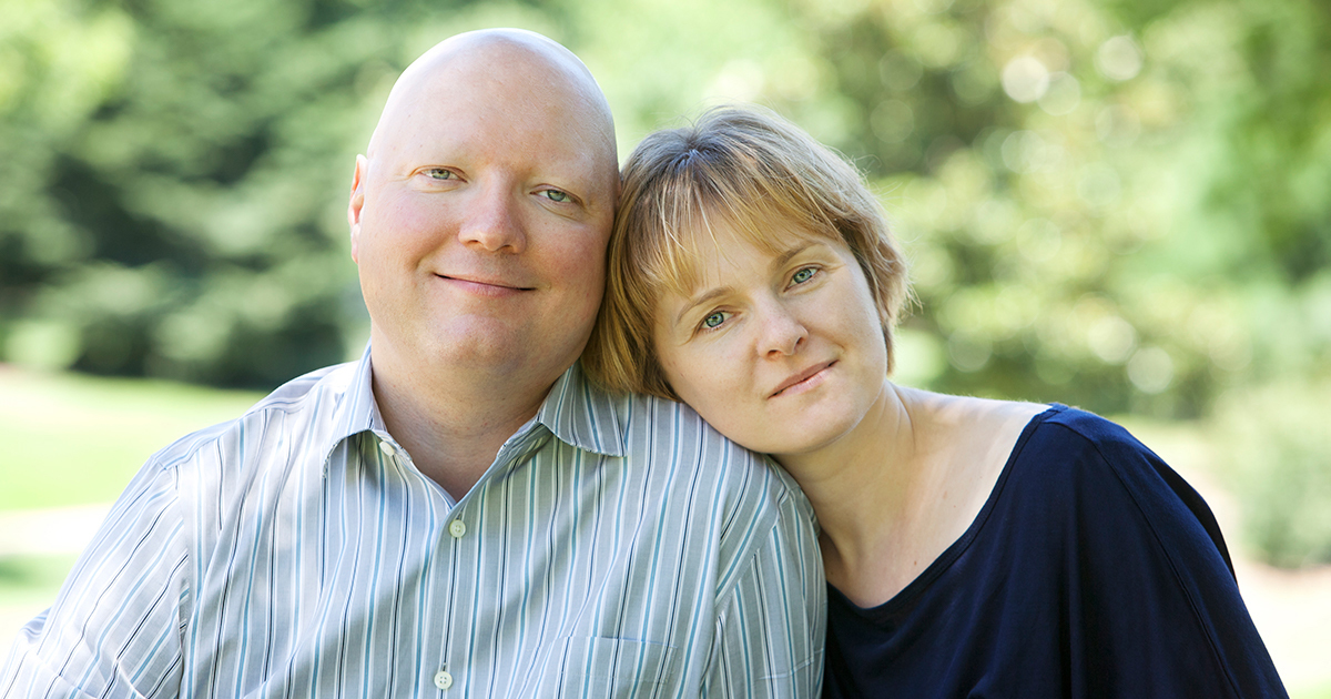 An older Caucasian couple sit next to each other, her head resting on his shoulder.