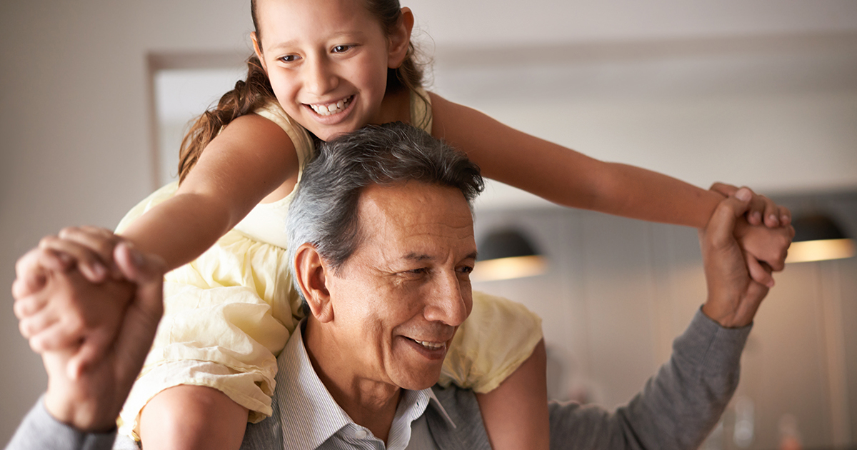 A man in a gray shirt standing indoors with his daughter seated on his shoulders.