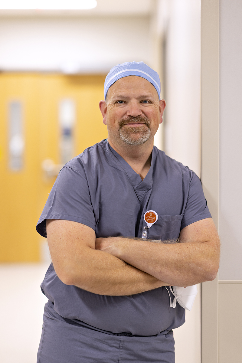 Karl Koenig, MD, surgeon in the Musculoskeletal Institute, stands in a hallway with arms crossed dressed in scrubs.