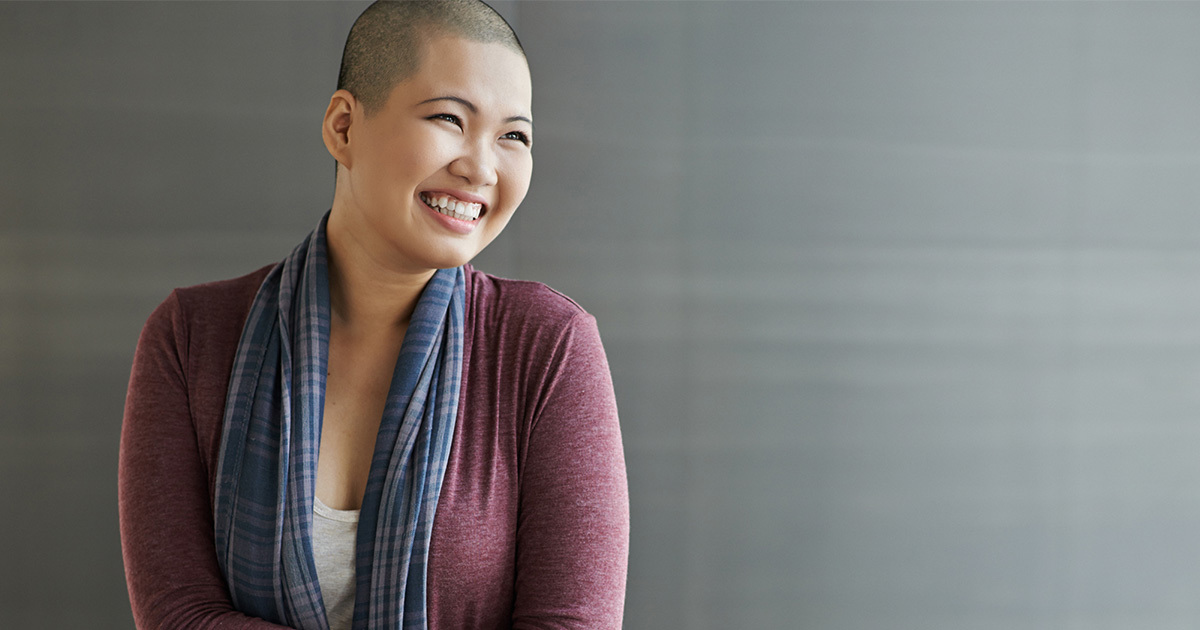 A young Asian woman with a shaved head is dressed in a burgundy sweater with a blue scarf and stands against a gray background.