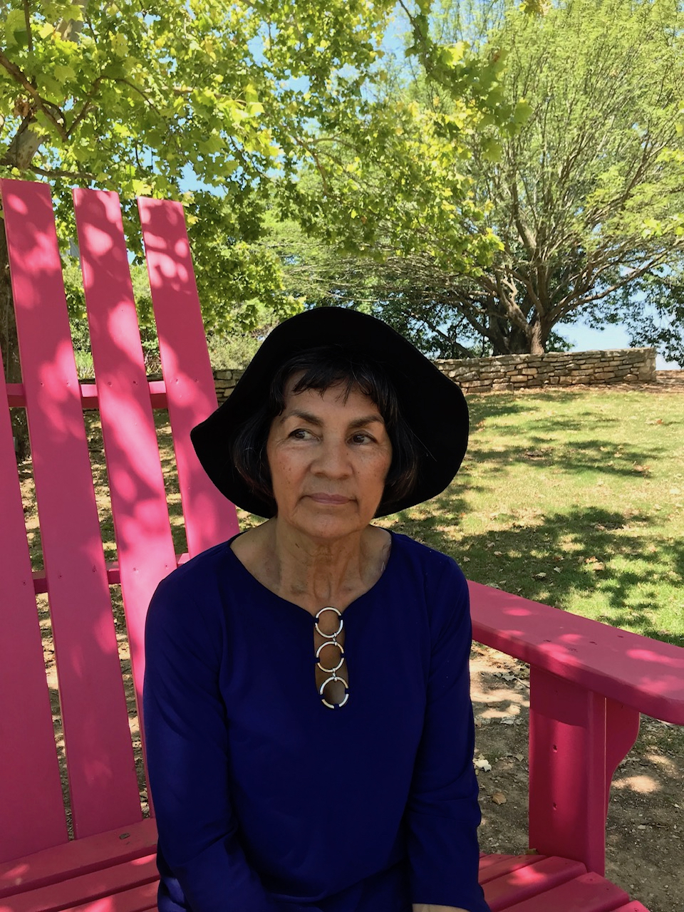 Mary, wearing a large black hat, sits in a large pink Adirondack chair in a park surrounded by trees.