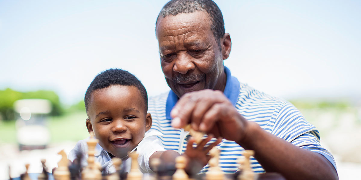 An elderly African-American man sits with his toddler grandson on his lap as they both reach toward game pieces on a chess board.