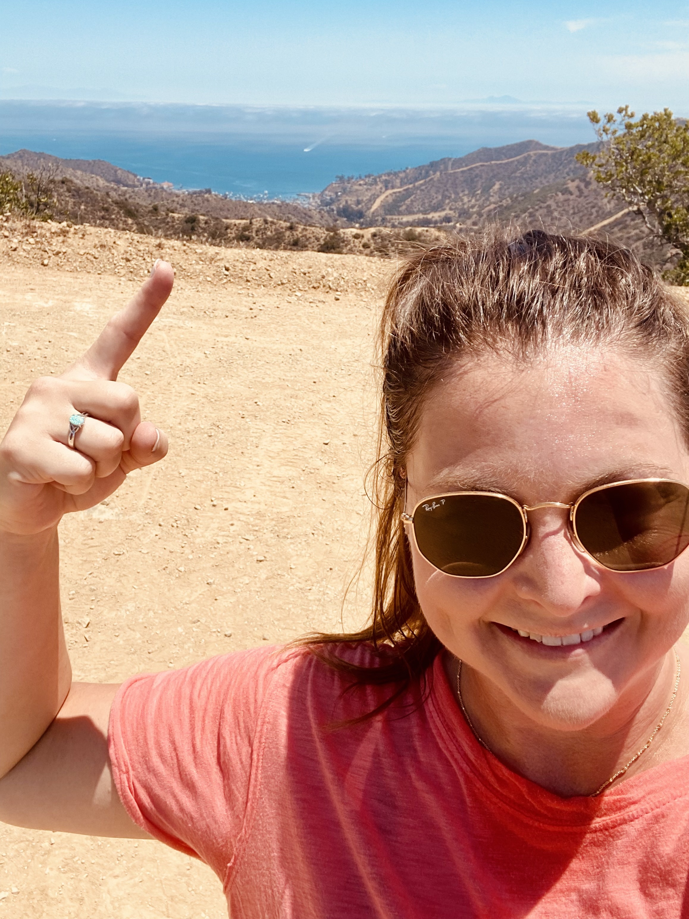 A woman with her hair up in a ponytail and sunglasses is taking a selfie and pointing at the view from the top of a peak on a hike.