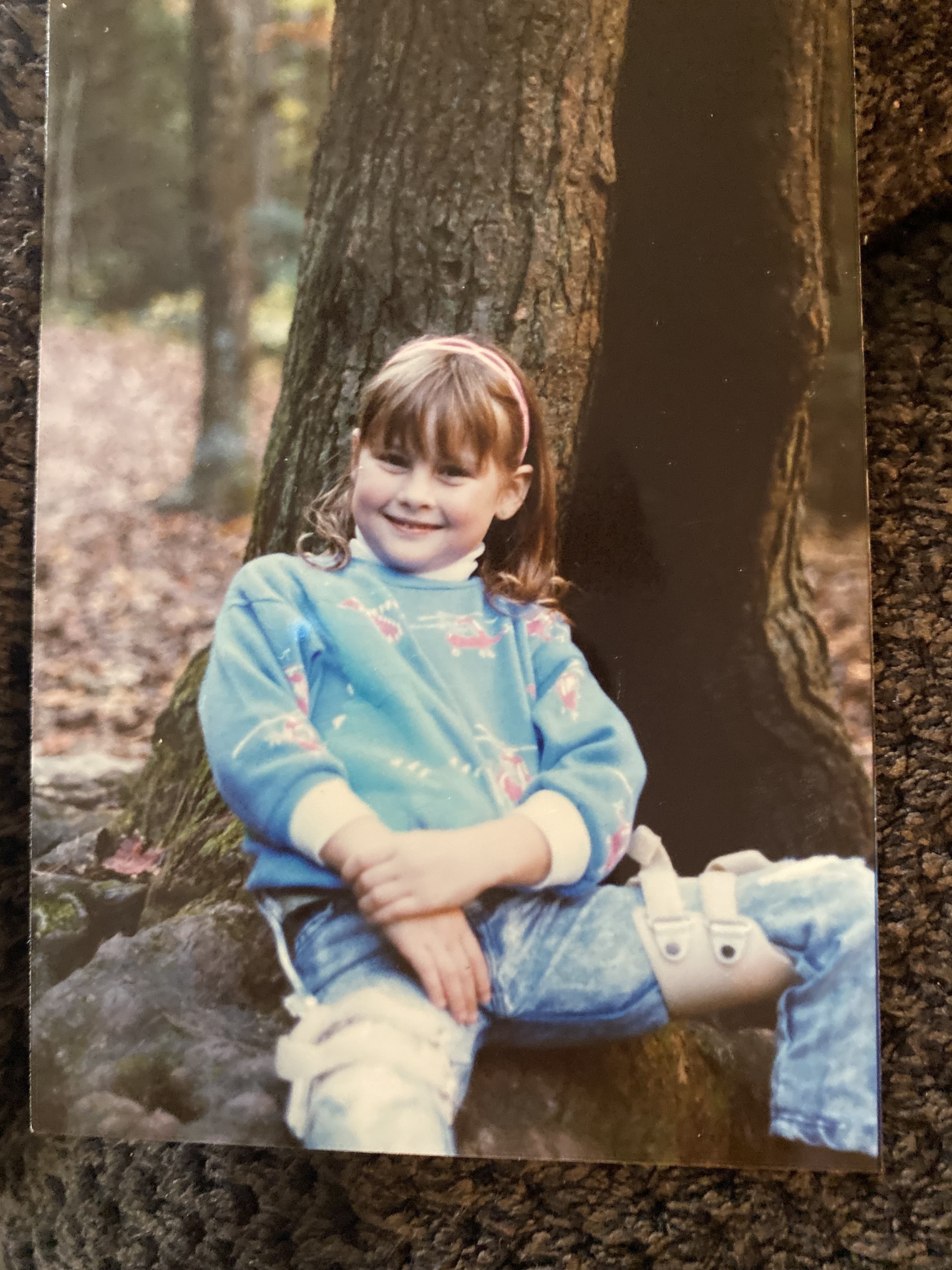 A young girl with bangs is wearing leg braces and sitting at the base of a tree trunk.