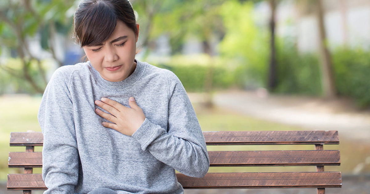 A young Asian woman sits on a park bench wearing a gray sweatshirt. She holds her hand to her chest and wears an expression of discomfort.