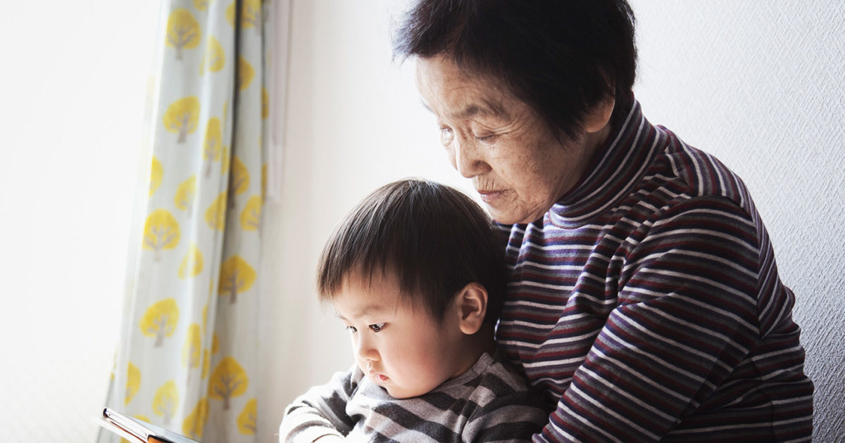 An Asian grandmother holds her grandson in her lap. She holds a mobile phone at which both of them are looking.