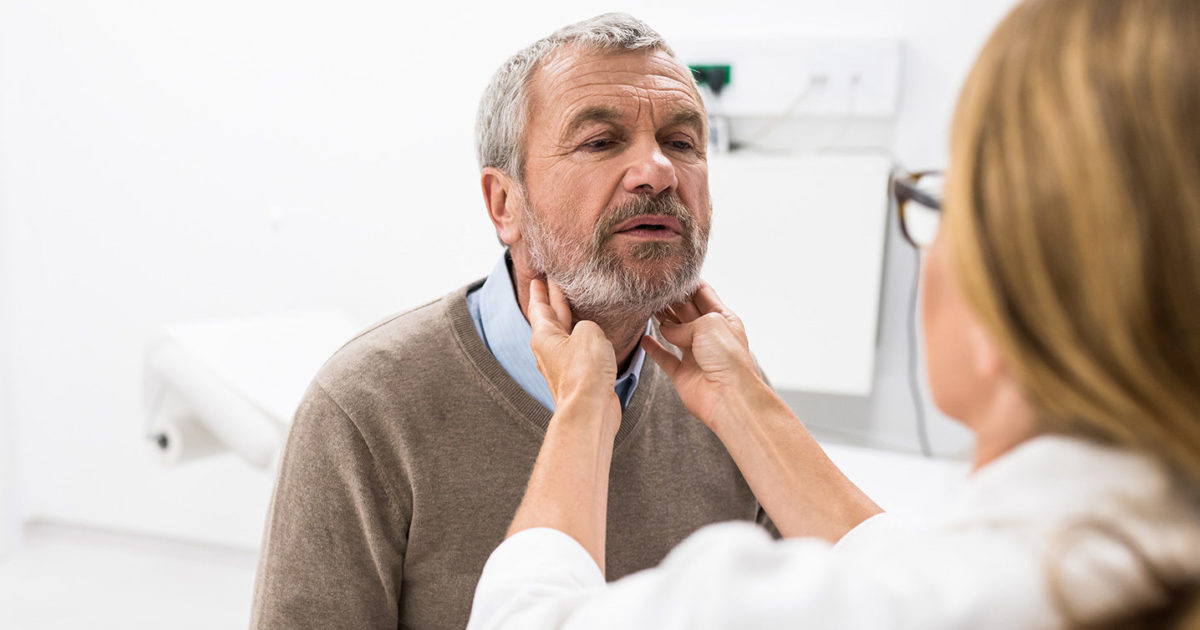 A man with gray hair and a light beard faces the camera while a female clinician places her fingers on his throat on either side of his chin.