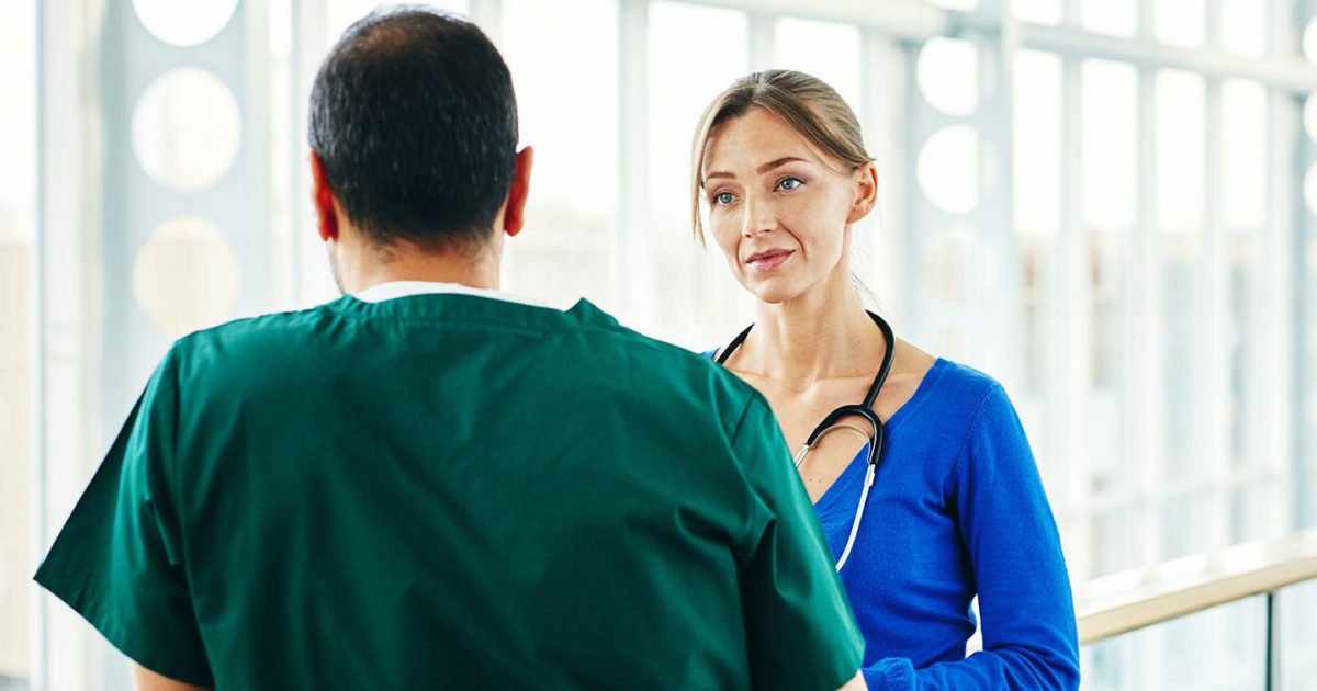 A man wearing medical scrubs stands with his back turned speaking to a woman who faces the camera, also wearing scrubs.