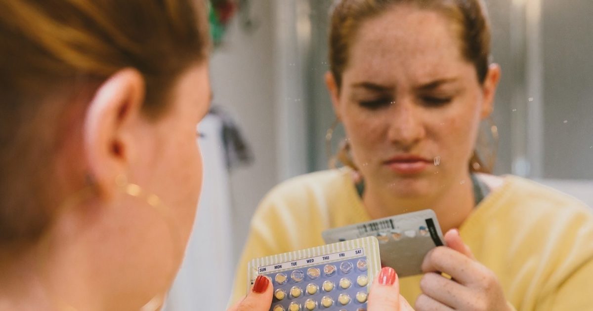 A young woman is seen in a bathroom mirror staring at a blister pack of birth control pills with a frowning, confused facial expression.