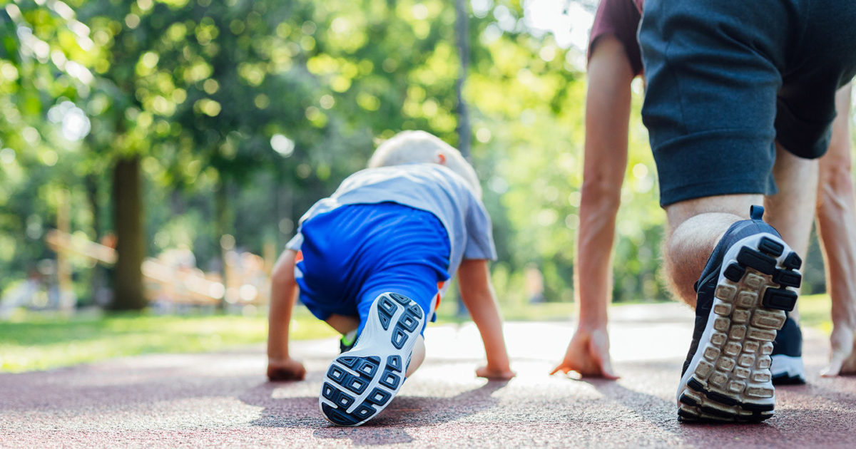 A young child squats on the ground in a runner's starting stance. Next to him, in the same position, is his father.