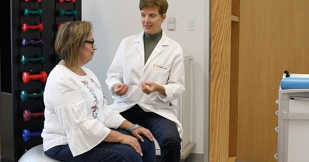 A UT Health Austin clinician sits speaking to a woman who sits on a Swiss ball.