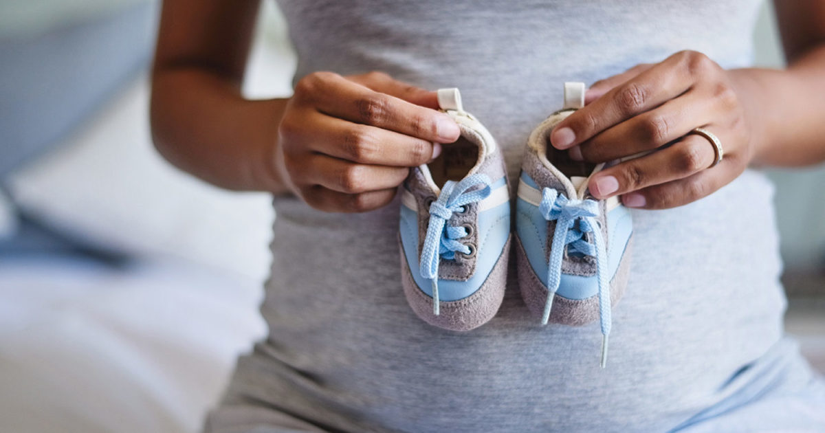 A pregnant woman holds a pair of baby shoes in front of her belly.