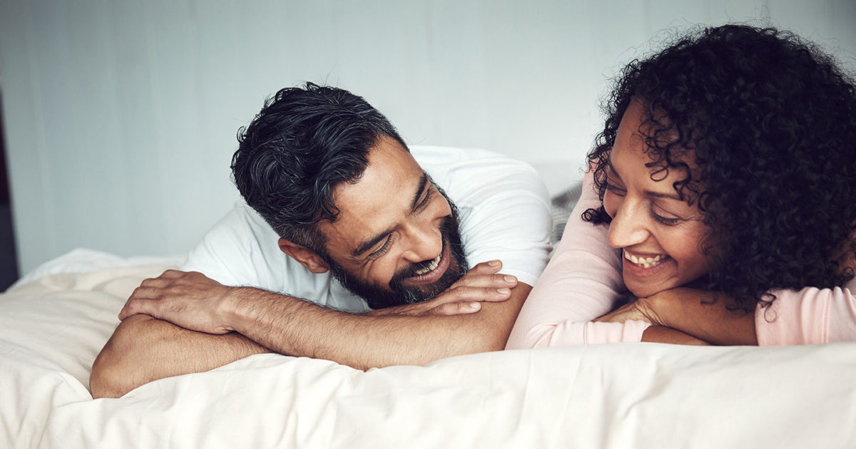 A male/female couple in their thirties lie on their stomachs next to one another in a bed. They look at each other and smile.