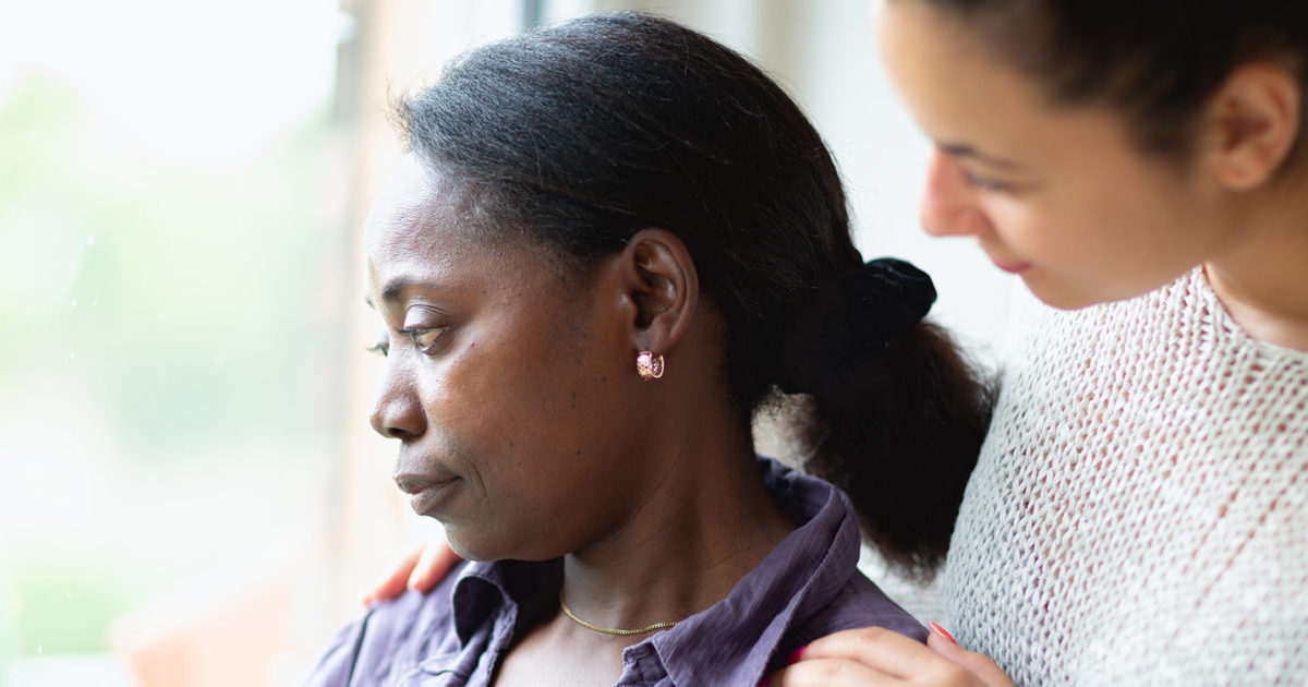 A middle-aged African-American woman stares out a window with a sad expression on her face. Behind her stands a younger woman who is placing her hands on the first woman's shoulders in a comforting expression.