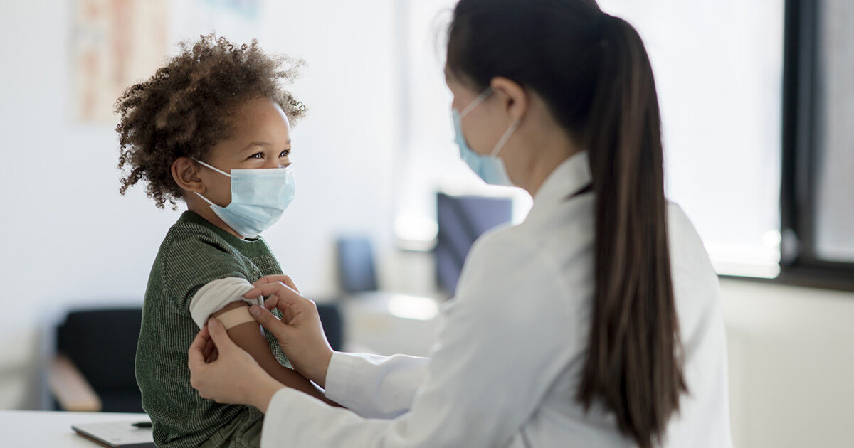 A young child wearing a forest green shirt is wearing a face mask and smiling at a provider who is putting a bandaid on their arm following a vaccination.