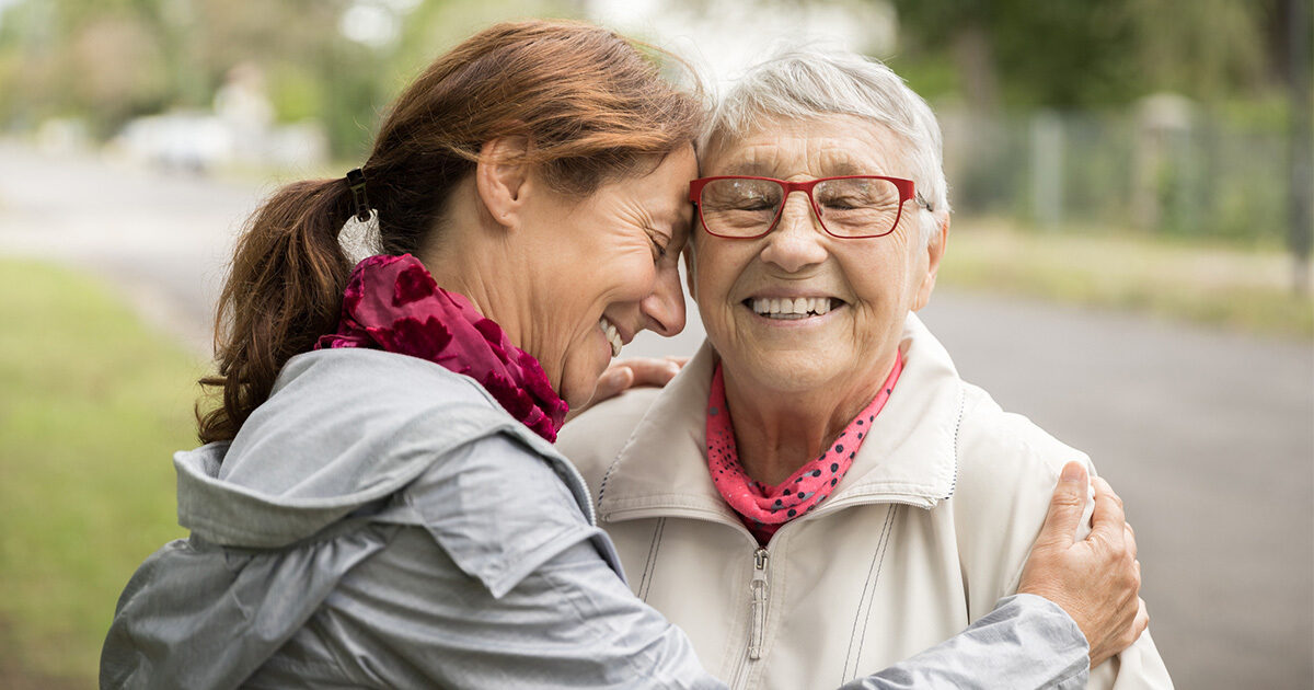 A middle aged woman with wavy red hair is hugging a smiling elderly woman with silver hair and red glasses.