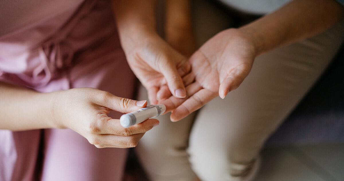 Close-up of a person in medical scrubs performing a blood glucose test on a patient's finger.