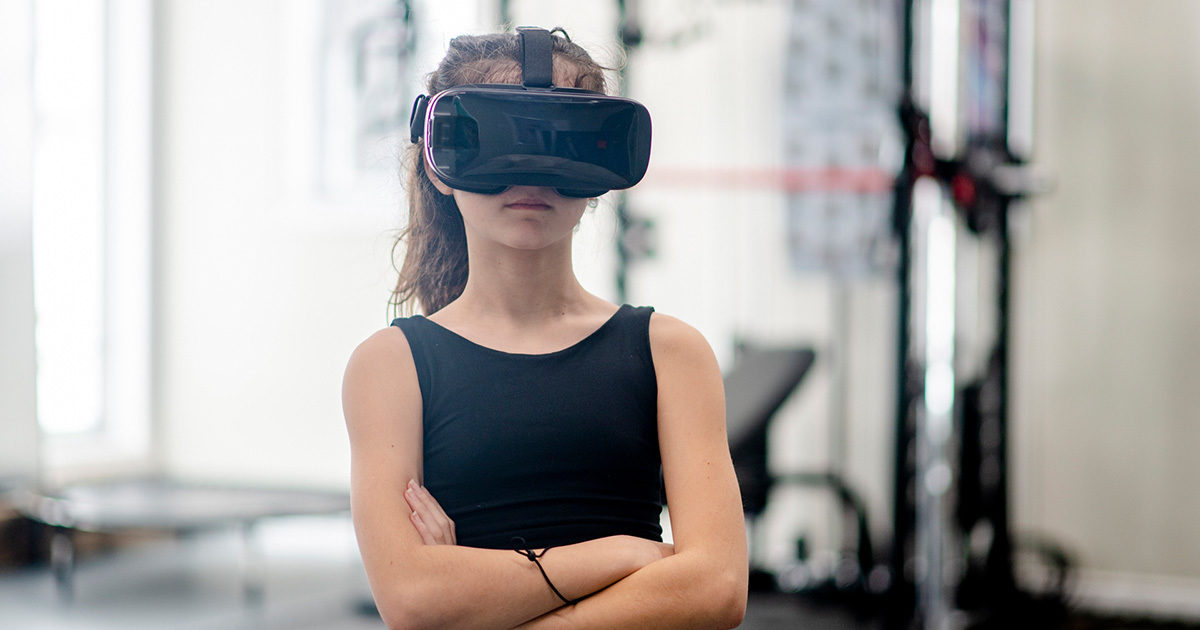 An adolescent girl with virtual goggles stands in a workout room with arms crossed across her waist.