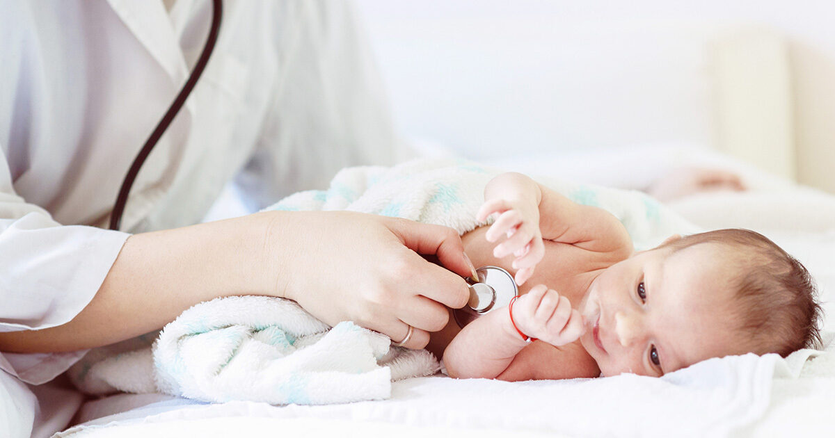 A small infant swaddled in a white blanket is laying on an exam table as a doctor holds a stethoscope to its chest.