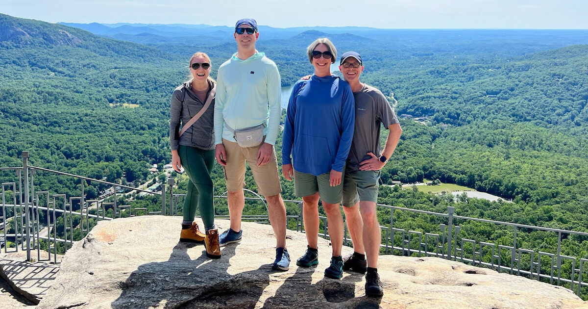 Joel Boehm, Katelyn Marak, and another couple stand on a boulder in hiking gear. Trees can be seen behind them far into the horizon.