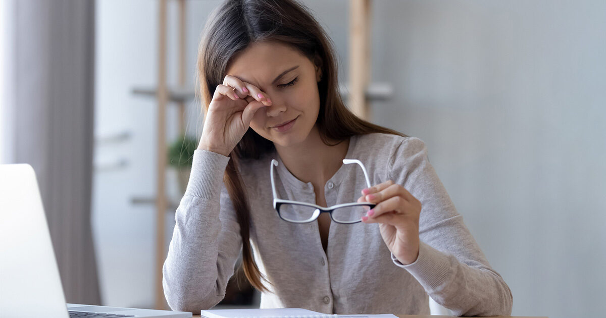 A young woman sits at her desk and is rubbing her eye with her left hand while holding her glasses in the other.