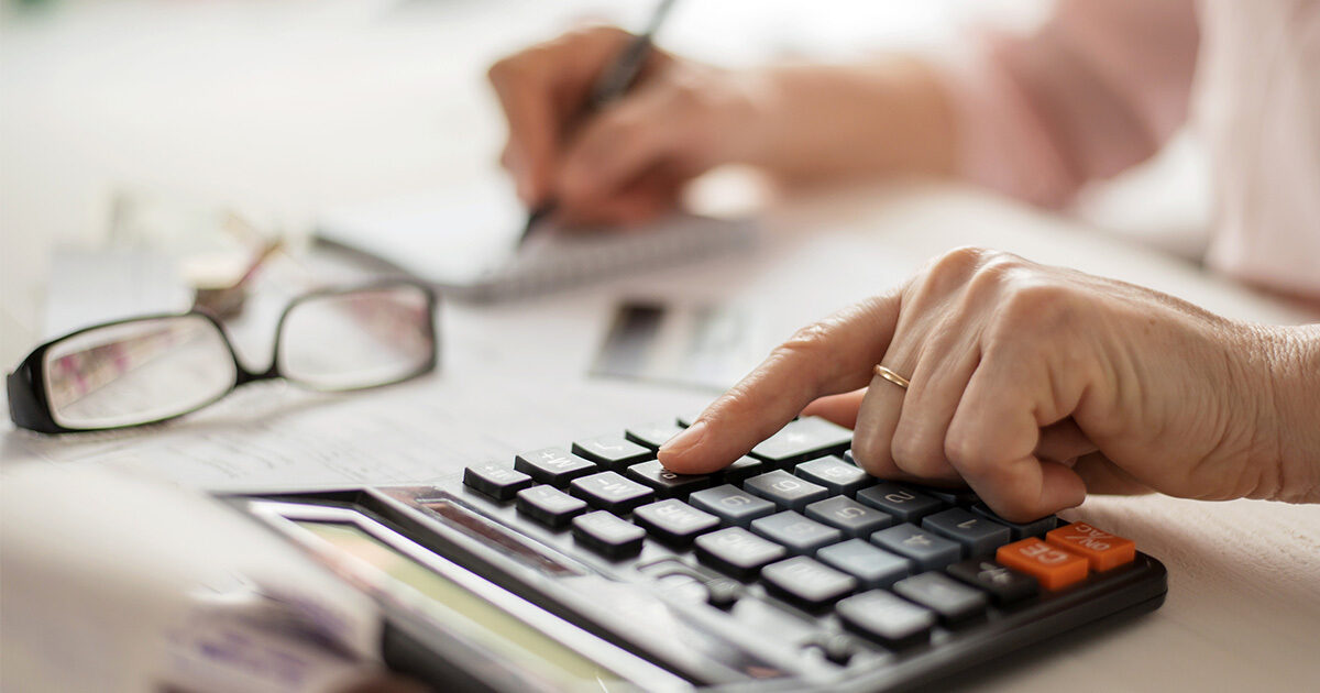 Close up of an older adult using a calculator with one hand and taking notes with their other hand. Her glasses sit on the table in front of her.