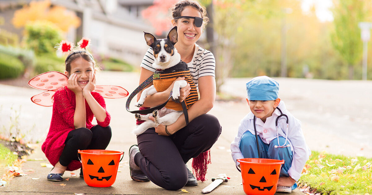 A mother and her two children are in Halloween costumes posing on the sidewalk with their jack-o-lantern candy buckets. The girl is a red insect, and the boy is a doctor in OR scrubs. The mother is a pirate, and she is holding onto their dog.