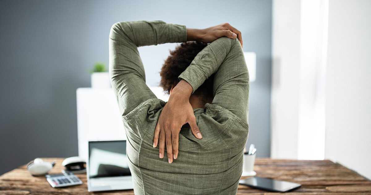 The backside of a woman wearing a forest green shirt is shown as she's sitting at a work desk and stretching. Her left arm reaches over her head and grabs her right elbow to stretch her triceps.