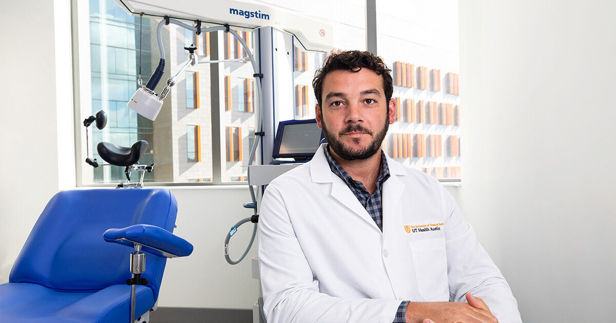 Dr. Nicholas Ortiz, who is wearing a white coat, sits in front of the transcranial magnetic stimulation machine, which holds a large magnet on an arm that extends over a blue chair.  The Health Discovery Building is seen in the windows behind him.