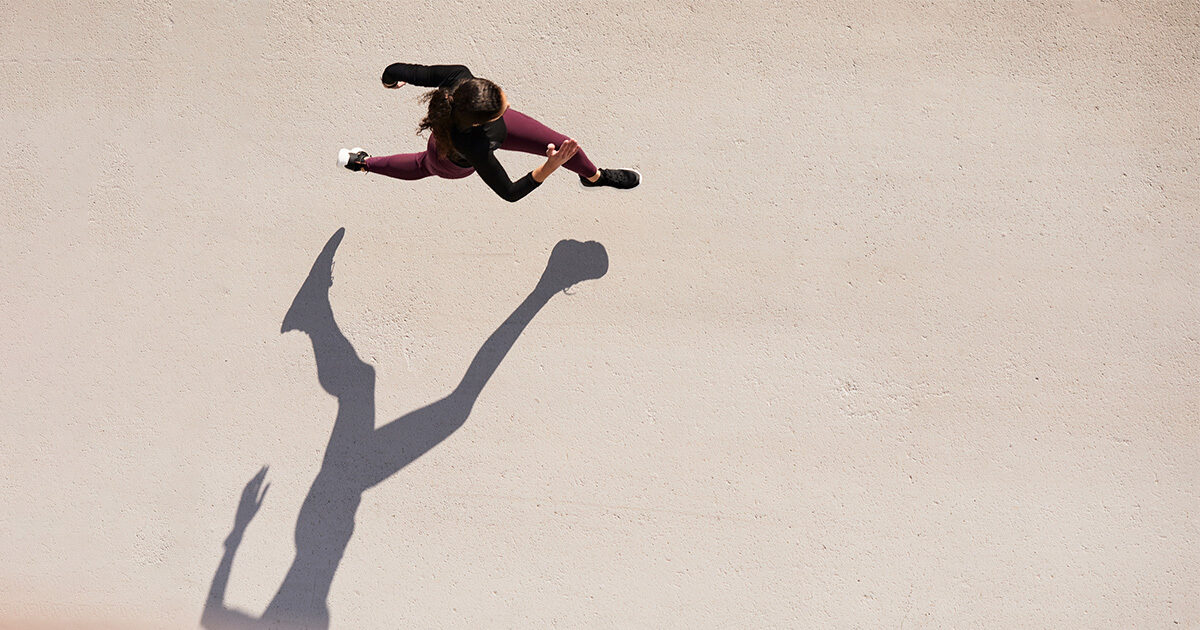 A young woman is seen sprinting from above in the bright sun.  She has dark brown hair, and is wearing a black top with purple leggings.  The sun casts a long shadow of her running.