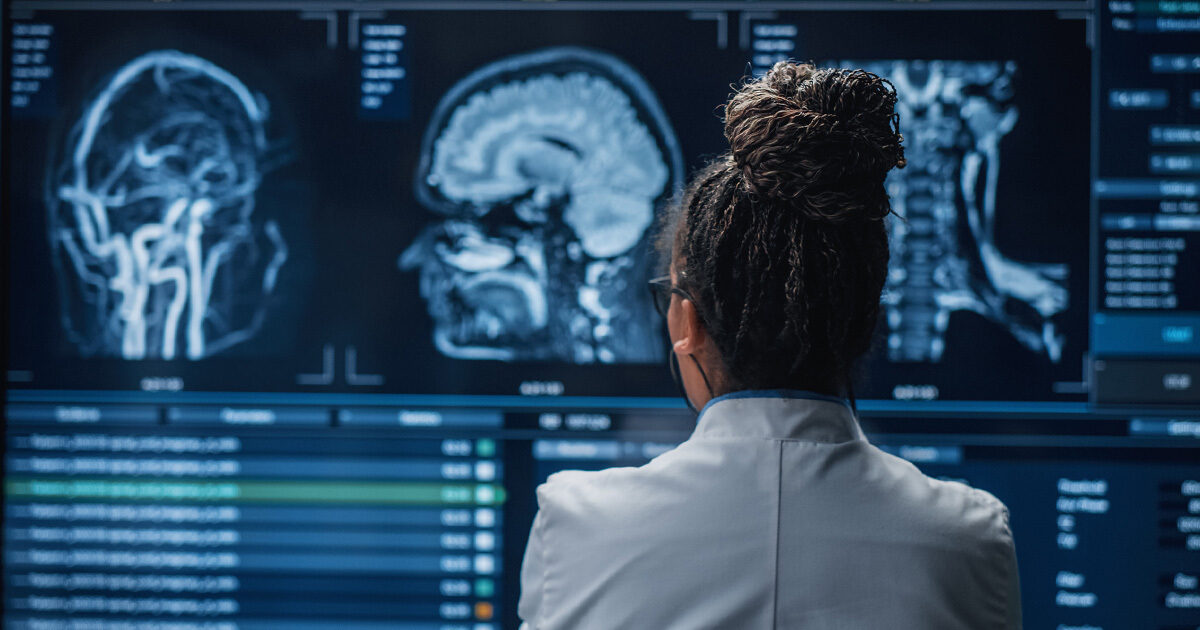 A physician in a white coat with her hair tied up in a bun is looking at three different brain scans in a dimly lit room.