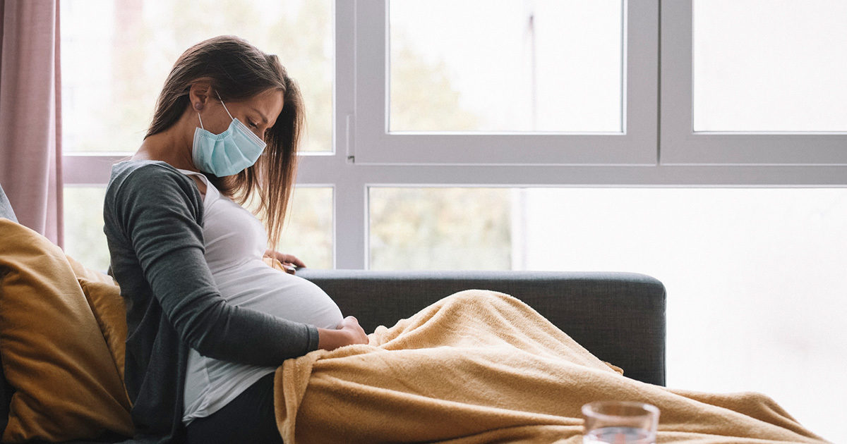 A young adult pregnant women, wearing a face mask, sits on her couch covered in a blanket.