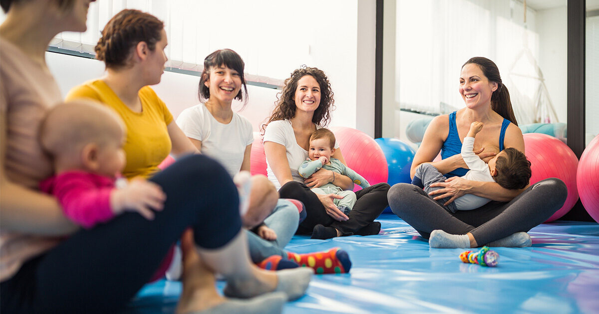 A group of women in an exercise class are seated on the ground holding their infant children.