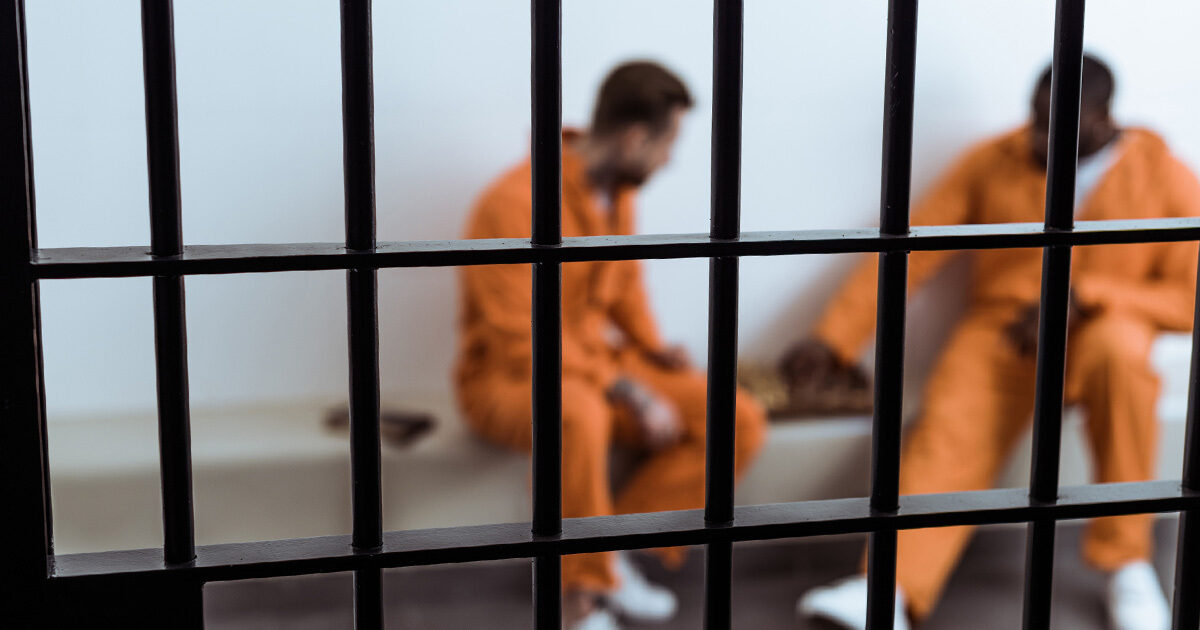 Two inmates are sitting on a bench wearing bright orange jumpsuits while playing a game of chess.