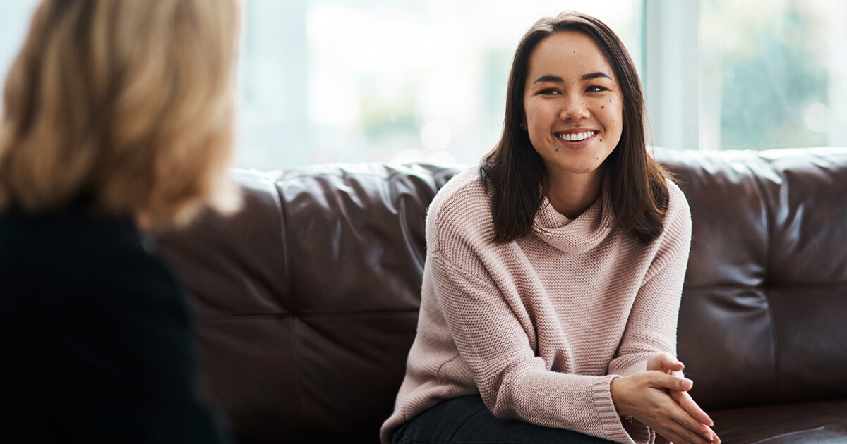 A young woman wearing a light pink turtleneck sweater is sitting on a dark brown leather couch with her hands clasped together, smiling at a therapist.