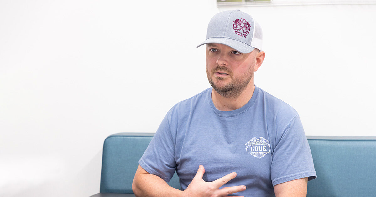 Tyler Cronin seated in an blue bench wearing a blue t-shirt and gray baseball cap.